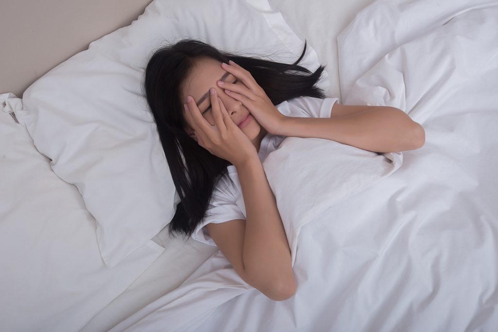 ผู้หญิงนอนอยู่บนเตียง พยายามใช้นิ้วนวดบริเวณหัวคิ้ว เพื่อคลายความตึงเครียดที่เป็นสาเหตุของอาการนอนไม่หลับ