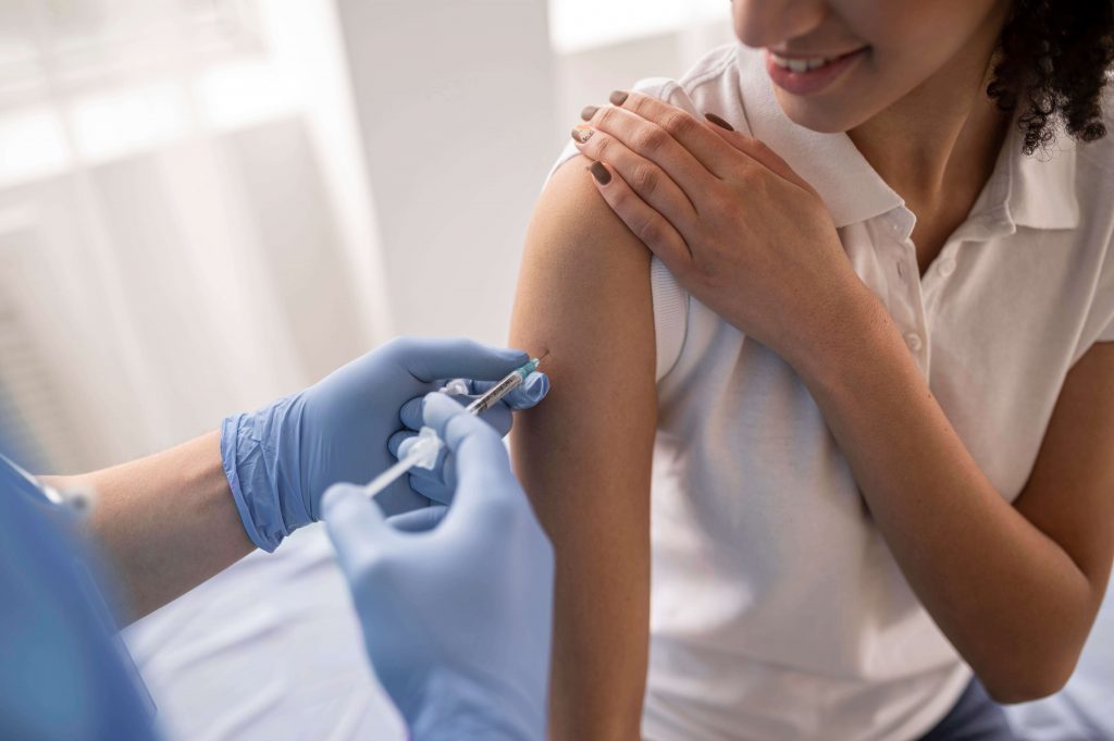 ผู้หญิงกำลังได้รับการฉีดวัคซีนที่ต้นแขนด้านขวา มีสีหน้ายิ้มแย้ม