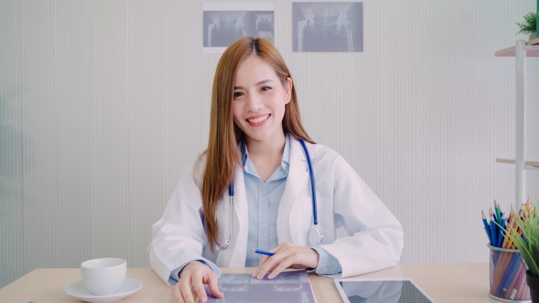 แพทย์หญิงกำลังนั่งทำงานในคลินิก พร้อมด้วยแท็บเล็ตสำหรับการจดบันทึก มองตรงมาที่กล้องด้วยรอยยิ้มสดใส