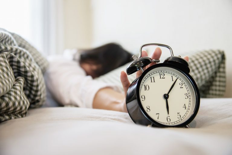 ควรนอนวันละกี่ชั่วโมงถึงจะสดชื่น? ผู้หญิงสะลึมสะลือเอื้อมคว้านาฬิกาปลุกที่ตั้งเตือนไว้ที่เวลา 6 โมงเช้าบนที่นอน