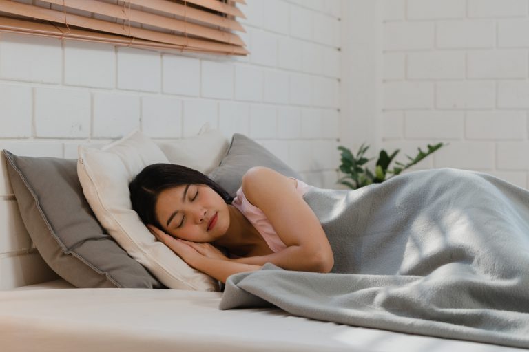 ผู้หญิงวัย 20+ กำลังนอนหลับลึกอยู่บนเตียงด้วยสีหน้ามีความสุข ท่ามกลางบรรยากาศที่เงียบสงบ ให้ความรู้สึกสบายตา